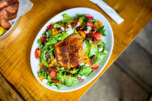 A vibrant salad topped with a seasoned fish fillet, mixed greens, tomatoes, pecans, and citrus segments on a white plate.
