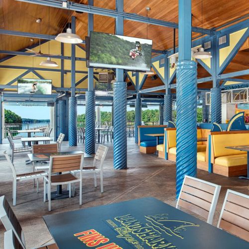 A bright, open restaurant with blue pillars, tables, and screens, overlooking a scenic waterfront view.