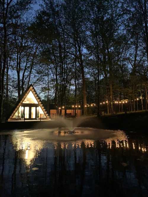 A triangular cabin illuminated at night, surrounded by trees and reflecting in a pond with a fountain.