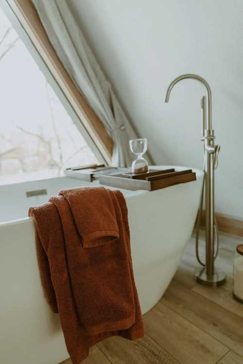 A cozy bathroom featuring a freestanding tub, a wooden tray, and a plush towel draped over the side. Natural light filters in.