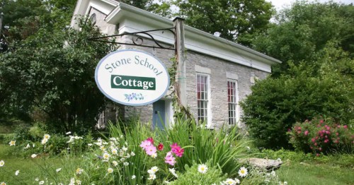 A stone cottage with a sign reading "Stone School Cottage," surrounded by colorful flowers and greenery.