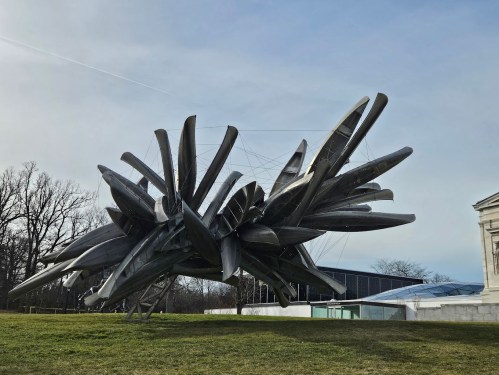 A large, abstract metallic sculpture with sharp, protruding shapes, set against a clear sky and grassy landscape.