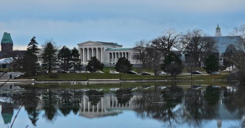 A serene view of a building with columns reflected in a calm lake, surrounded by trees and a cloudy sky.