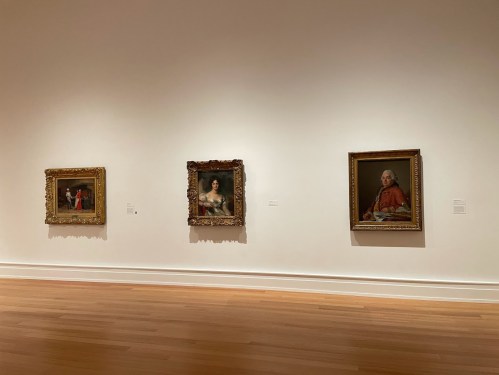 Three framed paintings hang on a white wall in a gallery, showcasing ornate frames and varied subjects.