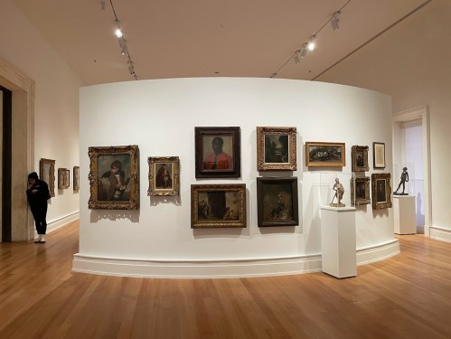 A gallery wall displaying various framed paintings and a sculpture, with a visitor observing the artwork.