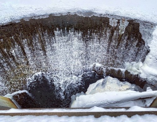 Aerial view of a circular water drainage structure with cascading water and snow around the edges.