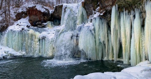 A frozen waterfall surrounded by snow-covered rocks and icy formations, with a serene pool at the base.