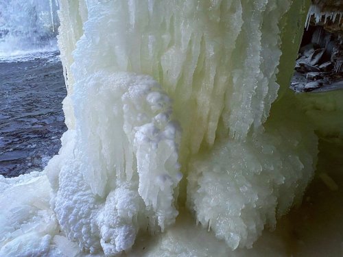 Close-up of frozen ice formations with intricate textures, glistening in natural light near a water source.