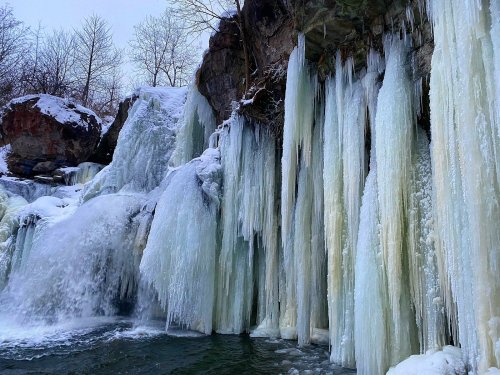 A frozen waterfall with icicles hanging from rocky cliffs, surrounded by snow-covered trees.