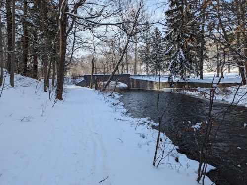 A snowy path alongside a river, with trees and a stone bridge in the background.