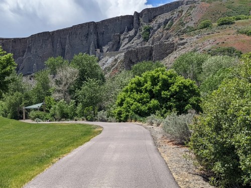 A winding path leads through lush greenery, with a rocky cliff and cloudy sky in the background.
