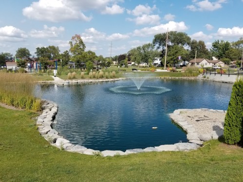 A serene pond with a fountain, surrounded by grass and trees, under a partly cloudy sky.