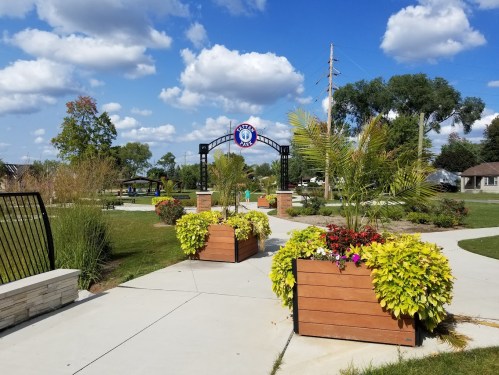 A park entrance with colorful flower planters, a blue sky, and fluffy clouds in the background.