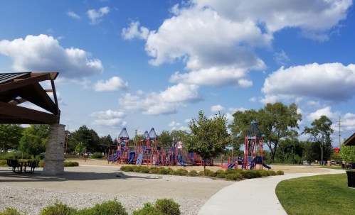 A colorful playground with slides and climbing structures, surrounded by green trees and a blue sky with fluffy clouds.