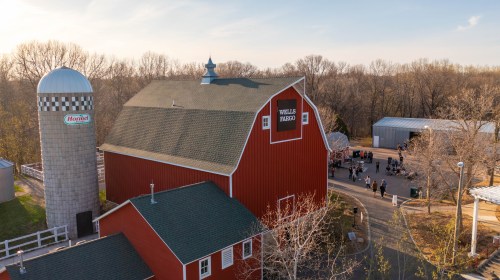 Aerial view of a red barn with a silo, surrounded by trees and people gathered outside in a rural setting.