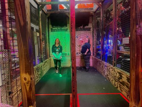 A woman stands in an axe-throwing arena, while a man prepares to throw in a colorful, illuminated setting.