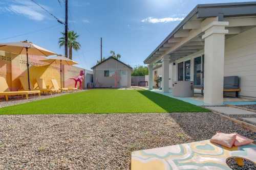 A sunny backyard with artificial grass, lounge chairs, umbrellas, and a small house in the background.