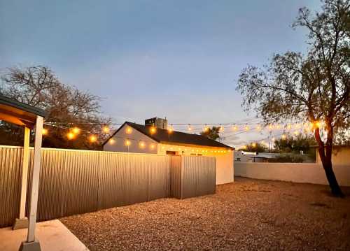 A backyard scene at dusk, featuring string lights overhead and a gravel area with a corrugated metal fence.