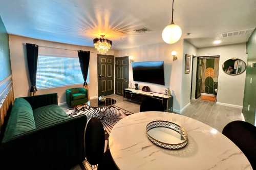 A stylish living room with a green sofa, round dining table, chandelier, and a large TV on the wall.