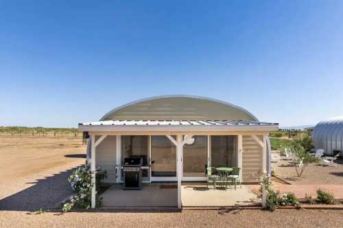 A modern, curved-roof house with a patio, grill, and outdoor seating in a rural landscape under a clear blue sky.