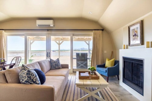 Cozy living room with a beige sofa, blue armchair, and large windows overlooking a beach view.