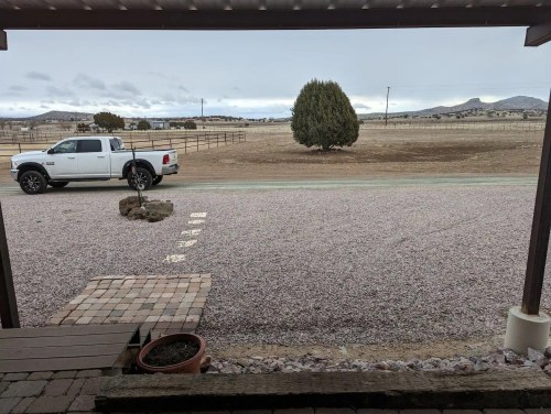 A gravel driveway with a white truck parked beside a large bush, overlooking a flat landscape under a cloudy sky.