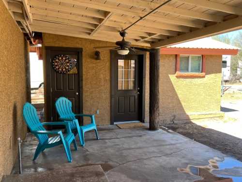 Two turquoise chairs on a concrete patio under a covered porch, with a dartboard and two doors in the background.