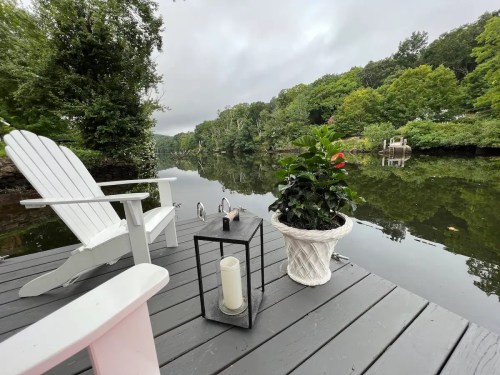 A serene lakeside scene featuring a white chair, a lantern, and a potted plant on a wooden dock surrounded by greenery.