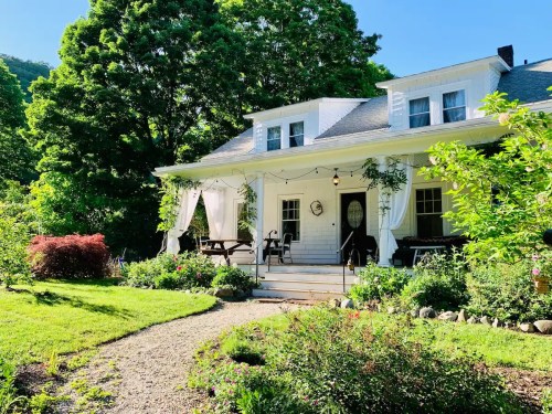 A charming white house with a porch, surrounded by lush greenery and colorful flowers on a sunny day.