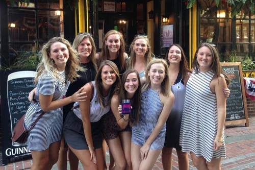 A group of nine women smiling together outside a restaurant, dressed in casual summer outfits.