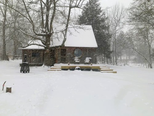 A cozy cabin surrounded by snow, with a wooden deck and chairs, under a snowy sky and bare trees.