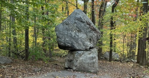 A large, balanced rock formation surrounded by colorful autumn trees in a forest setting.