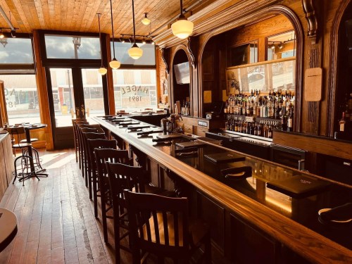A wooden bar with high stools, shelves of liquor, and large windows letting in natural light.