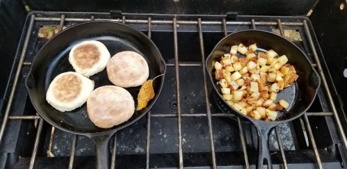 Two cast iron skillets on a grill: one with toasted English muffins and the other with diced, cooked potatoes.