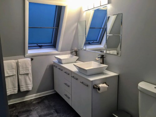 Modern bathroom with two vessel sinks, large mirrors, and skylights, featuring gray walls and white cabinetry.