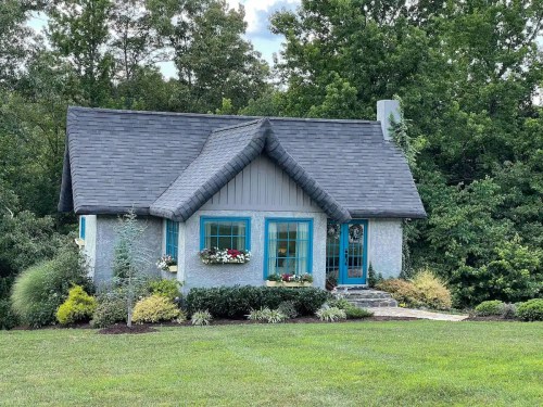 A charming cottage with blue trim surrounded by lush greenery and colorful flower beds on a sunny day.