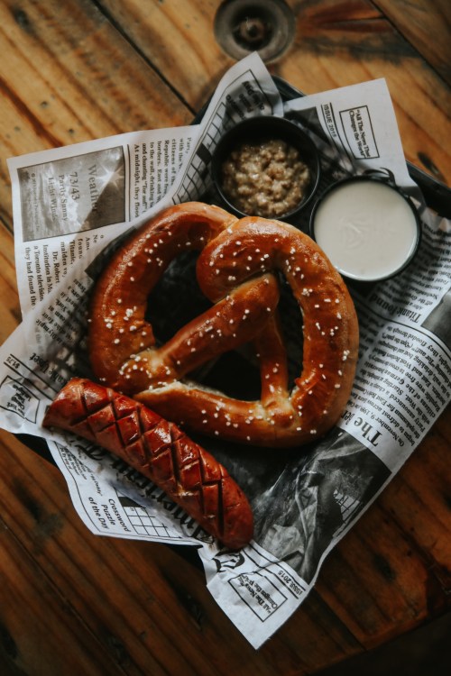 A basket with a large pretzel, a sausage, and dipping sauces, resting on newspaper.
