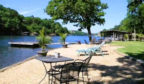 A serene lakeside scene with chairs, a table, and a tree, surrounded by water and greenery on a sunny day.