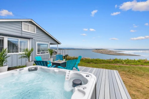 A hot tub on a deck overlooks a scenic view of water and a distant shore, with a cozy house in the background.