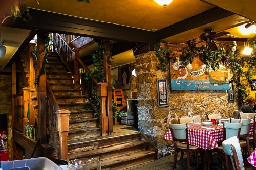 Cozy restaurant interior featuring wooden stairs, stone walls, and checkered tablecloths. Greenery decor adds charm.