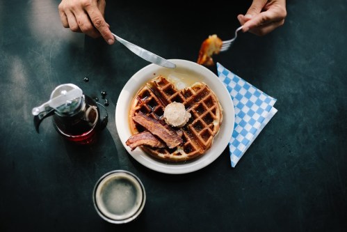 A plate of waffles topped with bacon and syrup, with a glass of drink and a napkin on a dark table.