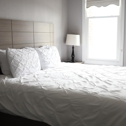 A neatly made bed with white textured bedding, a window with light curtains, and a bedside lamp in a bright room.