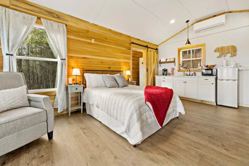 Cozy bedroom with wooden walls, a queen bed, armchair, and kitchenette, featuring soft lighting and a warm atmosphere.