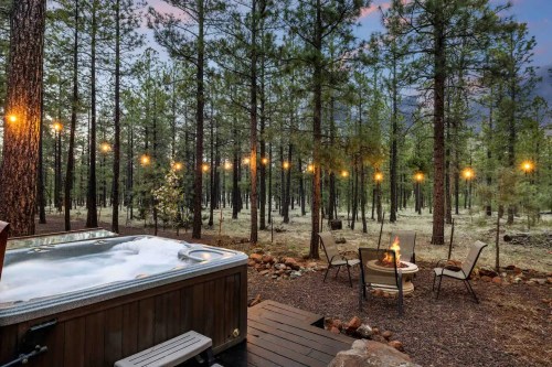 A hot tub beside a fire pit, surrounded by tall pine trees and string lights in a serene forest setting.