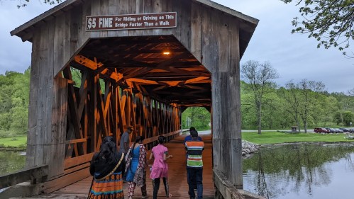 A group of people walking through a wooden covered bridge with a sign about fines for speeding. Lush greenery surrounds the area.