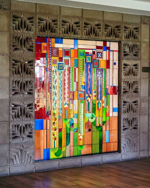 Colorful abstract stained glass artwork featuring geometric shapes and vibrant patterns, framed by textured stone walls.