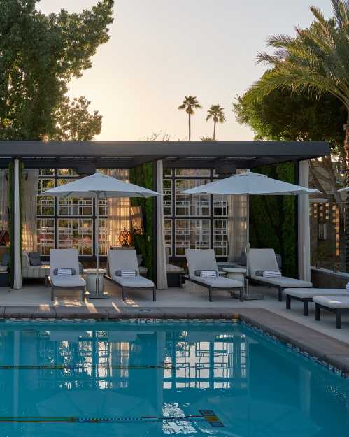 A serene poolside scene with lounge chairs, umbrellas, and palm trees under a sunset sky.