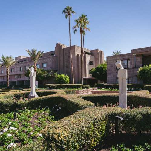 A landscaped garden with palm trees and geometric hedges, featuring stone sculptures and modern buildings in the background.