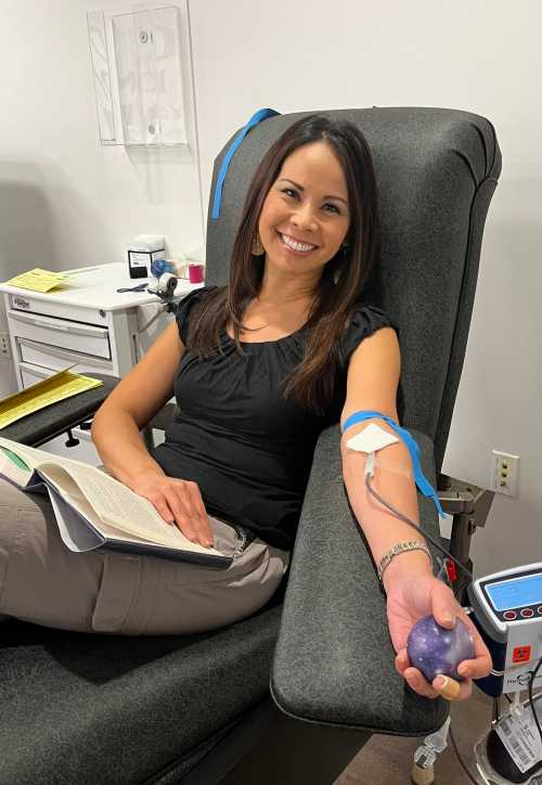 A woman sits in a donation chair, smiling while holding a book and a stress ball, with an IV in her arm.