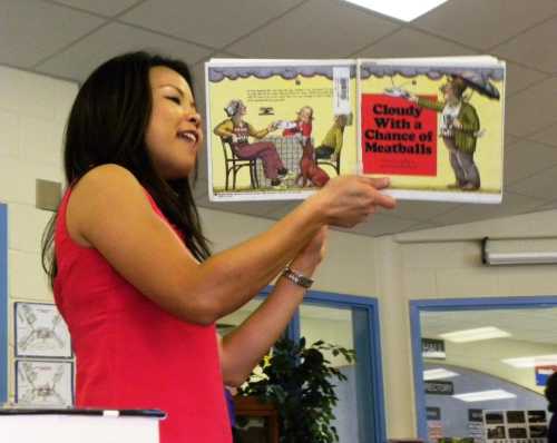 A woman in a pink top reads "Cloudy With a Chance of Meatballs" to an audience in a classroom setting.